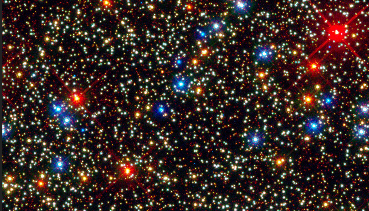 telescope photo showing hundreds of stars, most of them yellowish-white but some red and some blue