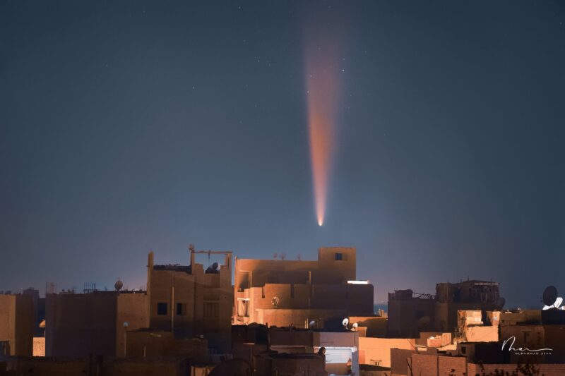 Comet with a pinkish tail above cream-colored buildings at night.