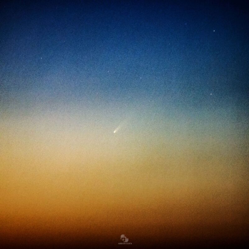 Rainbow-colored sky before dawn with a white comet with a tail going to the upper right.