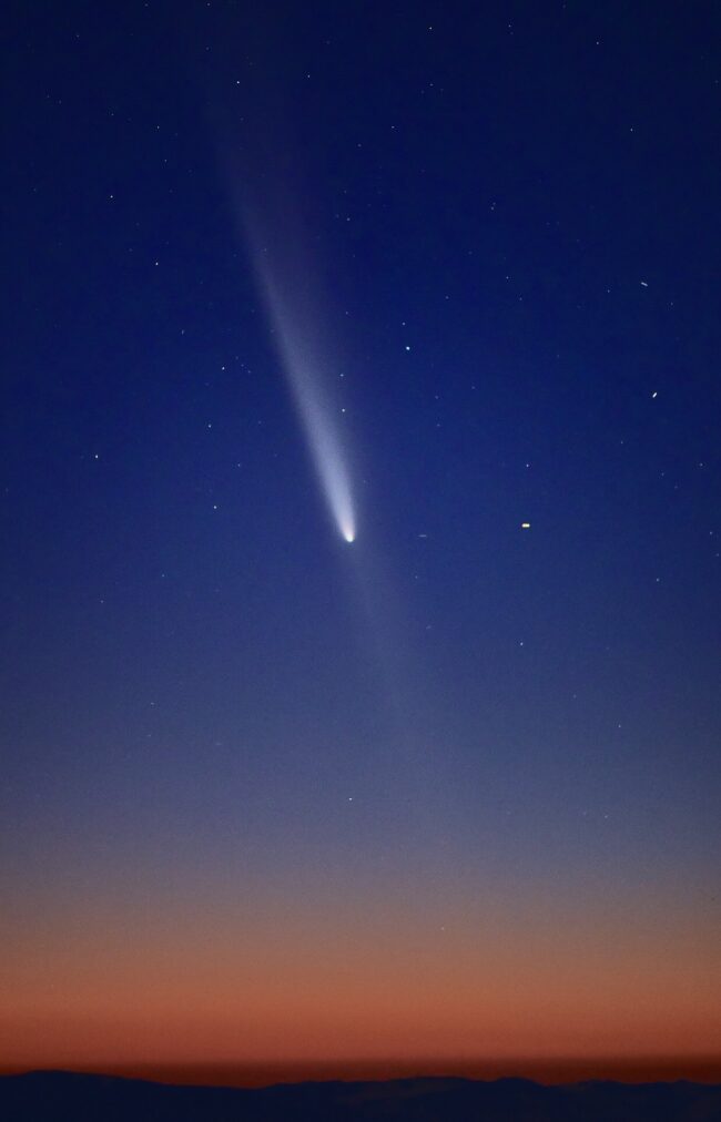 Red and blue sunset with a comet with a bright tail and a faint white line extending down from the head toward the ground.