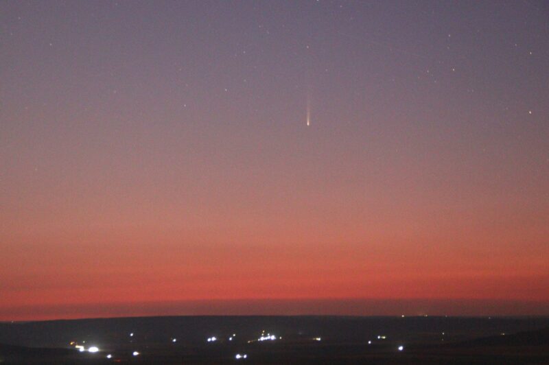 Pinkish blue sky with a comet above a city with lights.