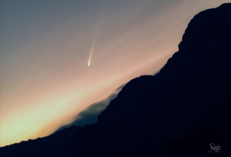 Dark hillside with lightening sky behind and a white comet with long tail above.