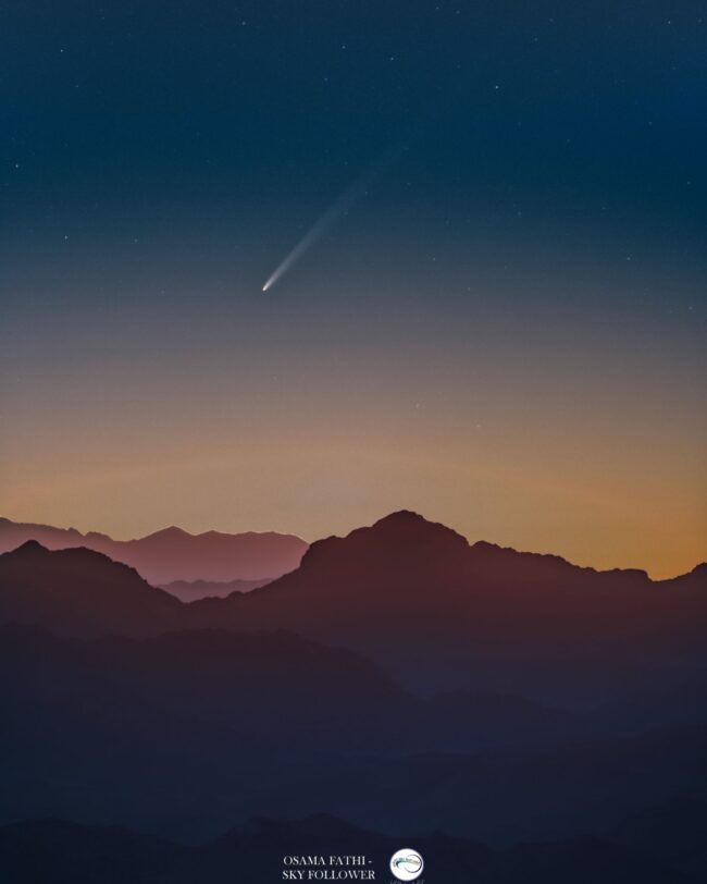 Comet over sepia-toned mountains in a sunrise sky.