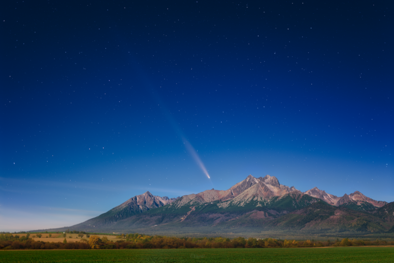 Dark blue sky at top and light blue at bottom. There are many small, white stars. A mountain in the background. There is a bright object with a fuzzy tail above the mountain.