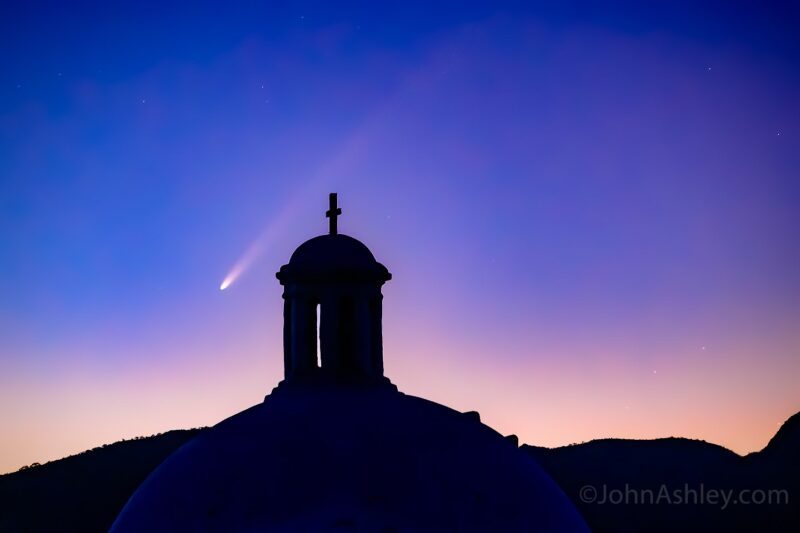 A mission with a cross on top silhouetted in a sunrise sky with a comet just behind.