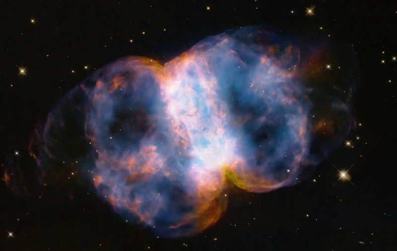 A Hubble image of the Little Dumbbell Nebula. The name comes from its shape, which is a two-lobed structure of colourful, mottled glowing gases that resemble a balloon that has been pinched around a middle waist. Like an inflating balloon, the lobes are expanding into space from a dying star seen as a white dot in the centre. Blistering ultraviolet radiation from the super-hot star is causing the gases to glow. The red colour is from nitrogen, and blue is from oxygen.