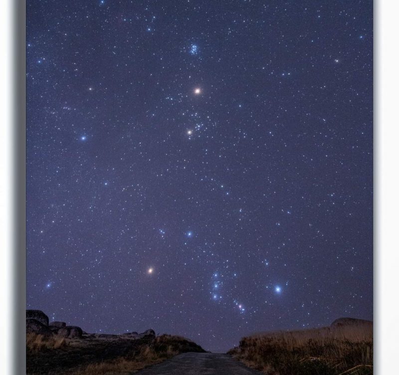 Starry sky with red Mars and Orion, Taurus, and the Pleiades over a rocky horizon.