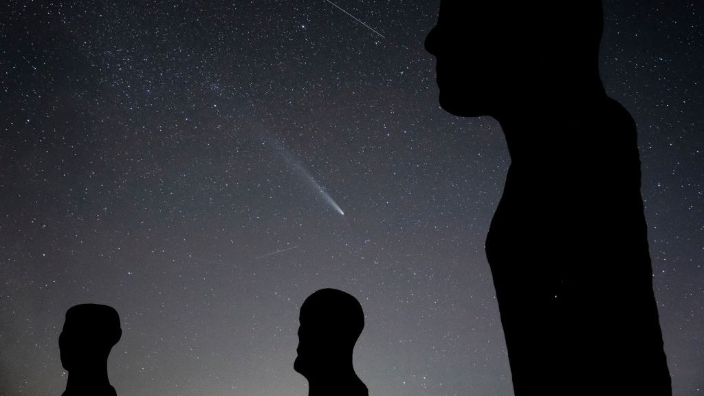 the long tail of a bright comet shines in a star-filled sky with figures silhouetted in the foreground.