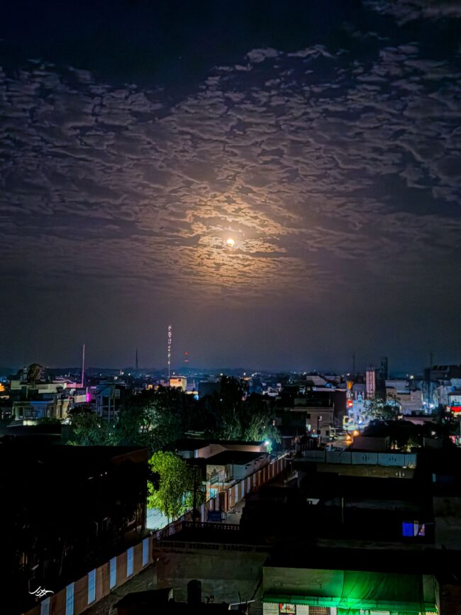 A moon peering between broken clouds and a city lit up below.