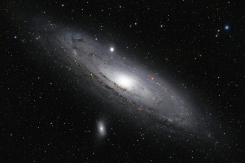 Large white disk with glowing center, bright reddish spots, dark lanes and thousands of foreground stars.