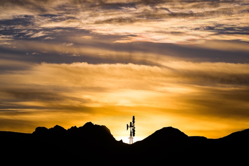 Golden sunrise with rays and clouds beyond dark hills and a silhouetted windmill.