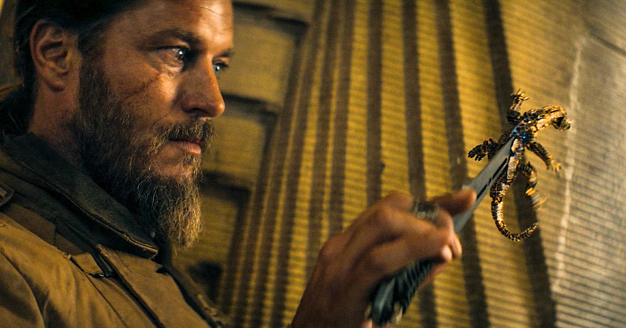 A bearded man holds a robotic lizard by the tip of his knife