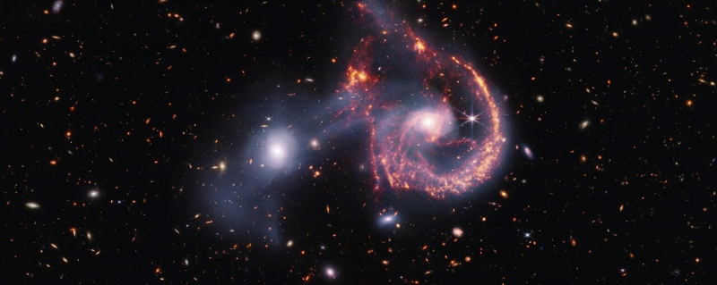 A pair of interacting galaxies. The larger of the two galaxies is slightly right of centre, and is composed of a hazy, bright, white center and a ring of gaseous filaments, which are different shades of red and orange. Toward the bottom left and bottom right of the ring are filaments of gas spiraling inward toward the core. At the top left of the ring is a noticeable gap, bordered by two large, orange pockets of dust and gas. The smaller galaxy is made of hazy and white gas and dust, which become more diffuse further away from its center. To this galaxy’s bottom left, there is a smaller, more diffuse gas cloud that wafts outward toward the edges of the image. Many red, orange, and white galaxies are spread throughout, with some being hazier in appearance and others having more defined spiral patterns.