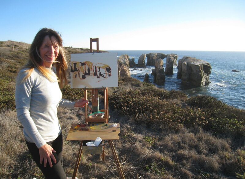 Smiling woman over a cliff, standing next to a painting. The sea is visible in the background. There are big rocks in a circle, in the water, close to the cliff.