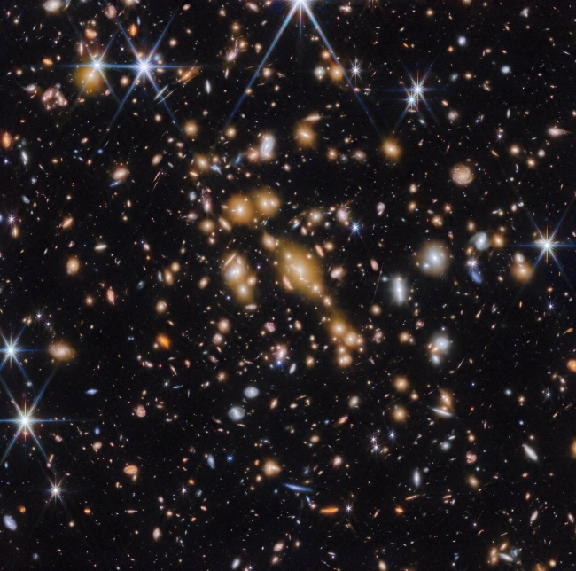 A field of galaxies on the black background of space. In the middle is a collection of dozens of yellowish galaxies that form a foreground galaxy cluster. Among them are distorted linear features, which mostly appear to follow invisible concentric circles curving around the centre of the image. The linear features are created when the light of a background galaxy is bent and magnified through gravitational lensing. A variety of brightly coloured, red and blue galaxies of various shapes are scattered across the image, making it feel densely populated.