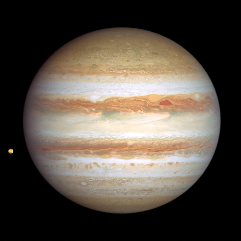 Jupiter is banded with stripes of brownish orange, light grey, soft yellow, and shades of cream, with many large storms and small white clouds punctuating the planet. At upper right of centre, a pair of storms appear next to each other: a deep-red, triangle-shaped cyclone and a reddish anticyclone. Toward the far-left edge of this view is Jupiter’s tiny orange-coloured moon Io.