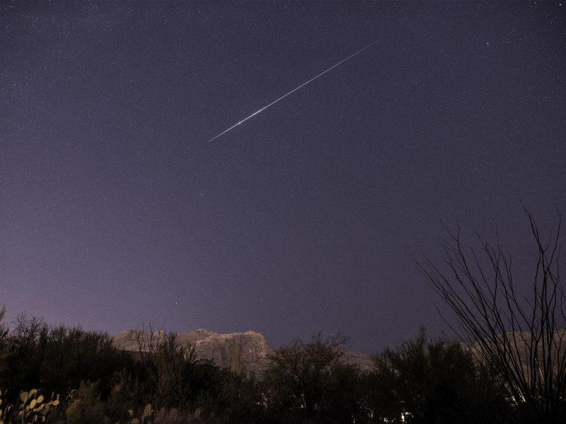 A purple-dark sky above a mountainous crag and bushes, with a single long streaking meteor in the sky.