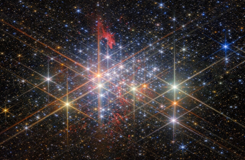 A dense cluster of bright stars, each with six large and two small diffraction spikes, due to the telescope’s optics. They have a variety of sizes depending on their brightness and distance from us in the cluster, and different colours reflecting different types of star. Patches of billowing red gas can be seen in and around the cluster, lit up by the stars. Small stars in the cluster blend into a background of distant stars and galaxies on black.