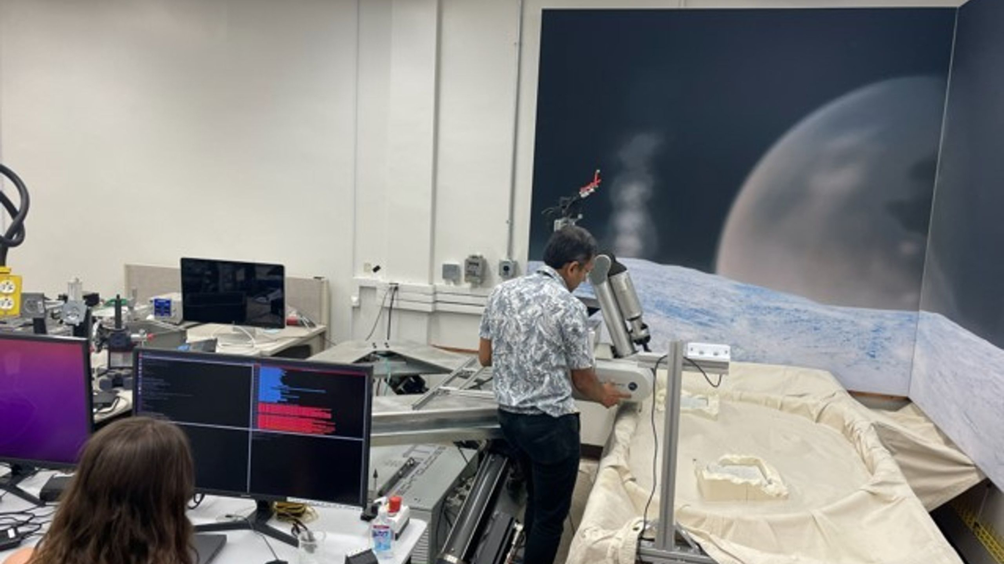 a man operates a robotic arm in a laboratory, with a large poster of a planet on the wall