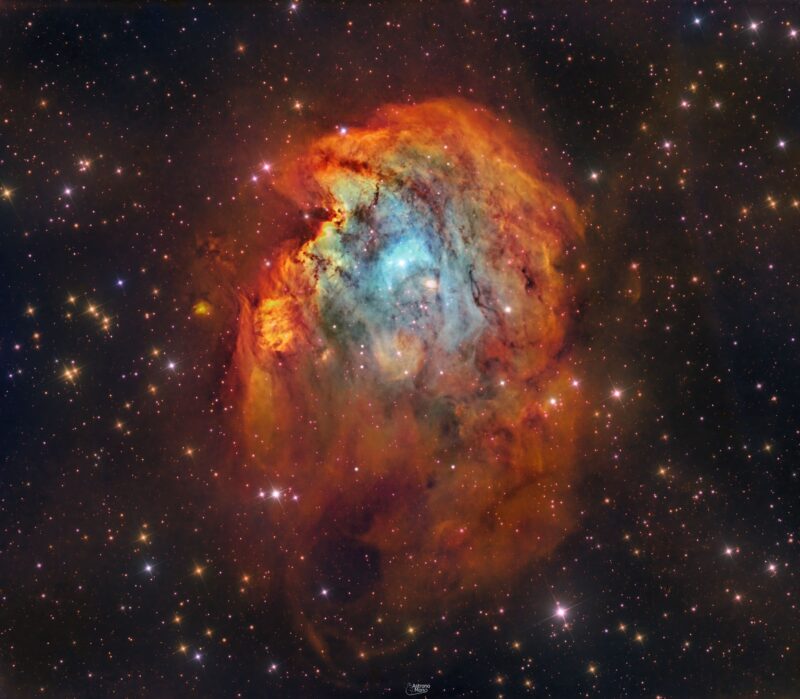 Large nebulous patch with bright orange and blue swirls, dark lanes and numerous foreground stars.