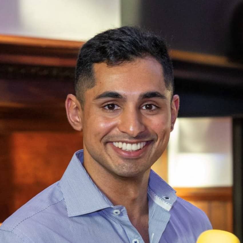 A young man with short black hair, wearing a light blue shirt, smiles at the camera