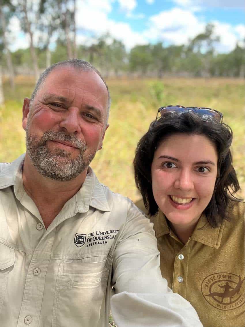 A man and young woman pose for a selfie. He has short grey hair and a beard, she has short brown hair. They are standing out in the bush.