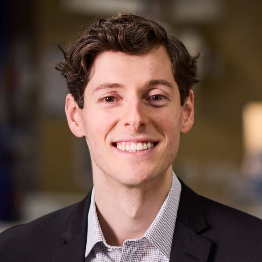 A young man with short brown hair, wearing a dark grey suit and white shirt, smiles at the camera