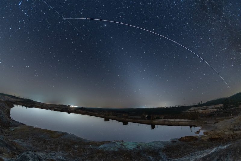 Dark starry sky with a white pyramid of light leaning to the left with an arc of light going across the sky, reflecting in a pond.