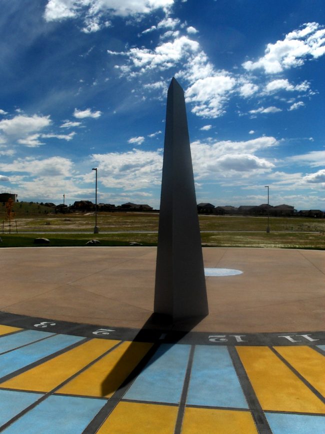 High black obelisk with shadow cast on radial blue and yellow checkerboard with times on it.