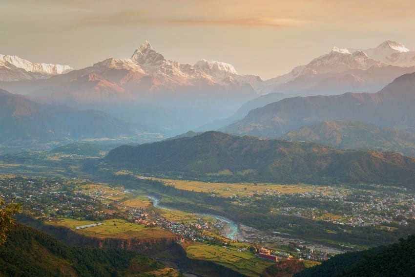 A view of distant snow-capped mountains and green vegetation and river surrounded by a village in the foreground