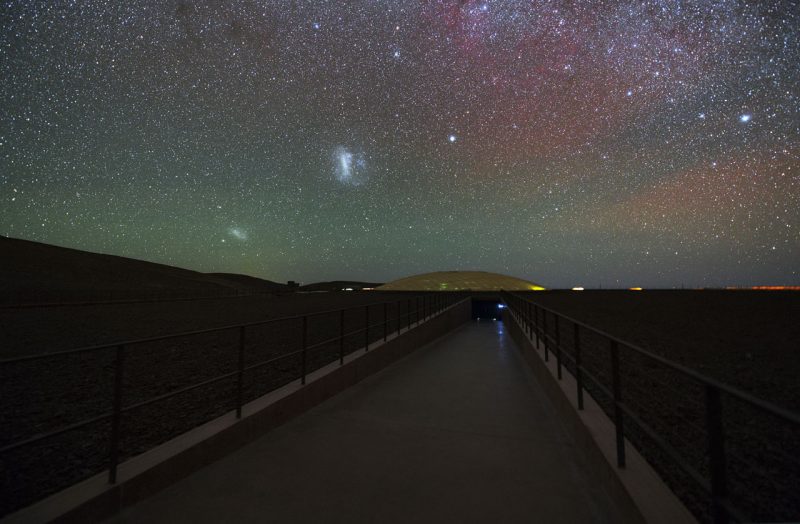 Starry sky with a large fuzzy patch and a smaller fuzzy patch to lower left above a road.