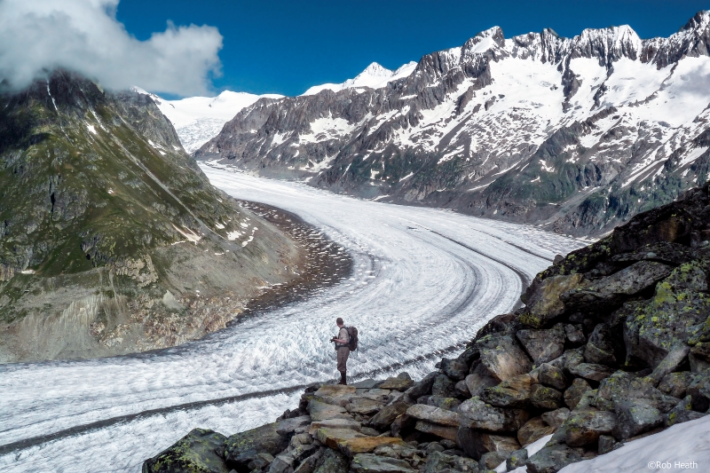 A wide icy stretch curves sharply through a rocky valley, with snow-covered peaks on the right. A man in hiking gear in the foreground looks at the glacier.