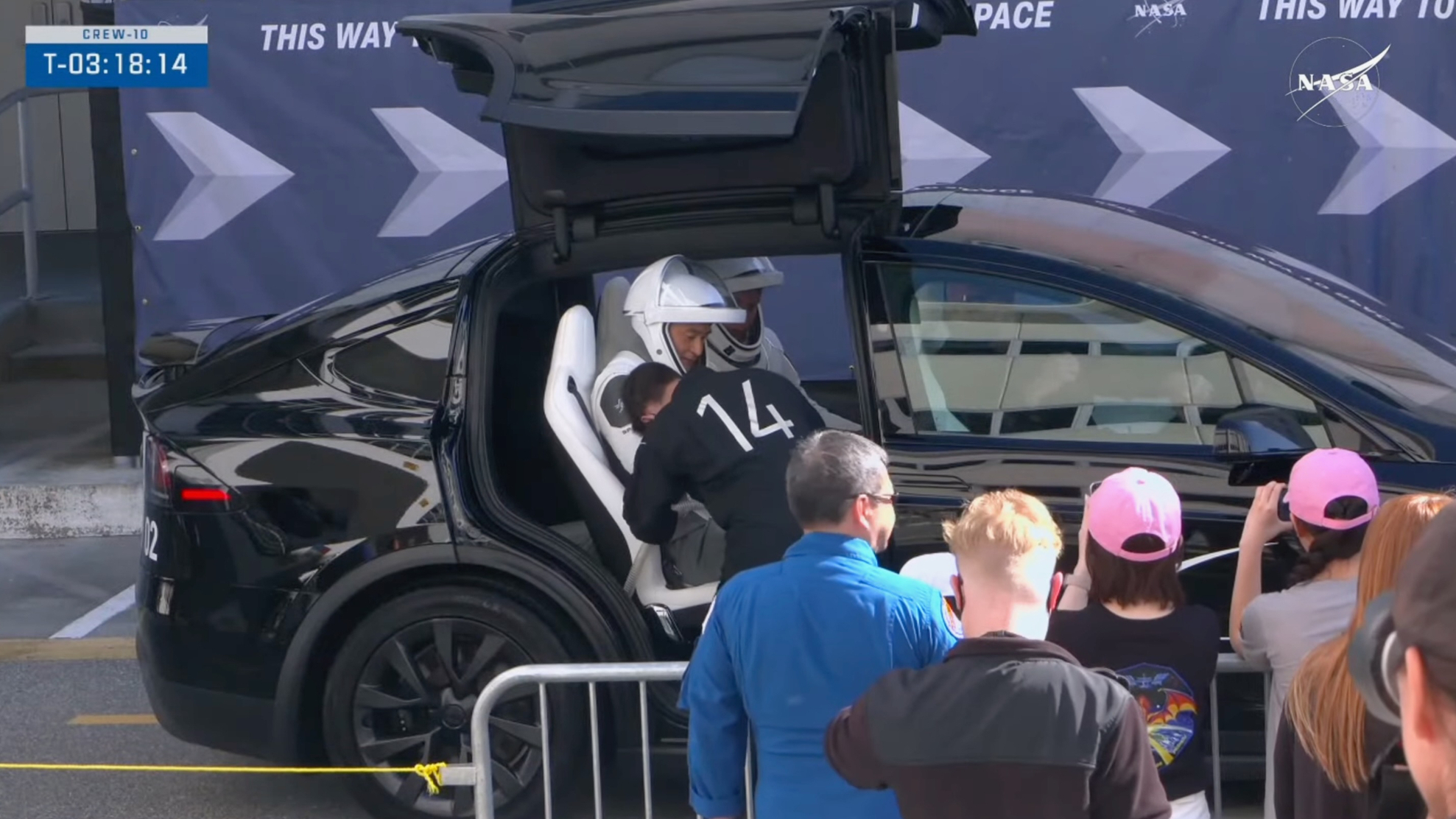 a man in a black jumpsuit helps an Cosmonaut in a white spacesuit get situated inside a black car, while Many people look on