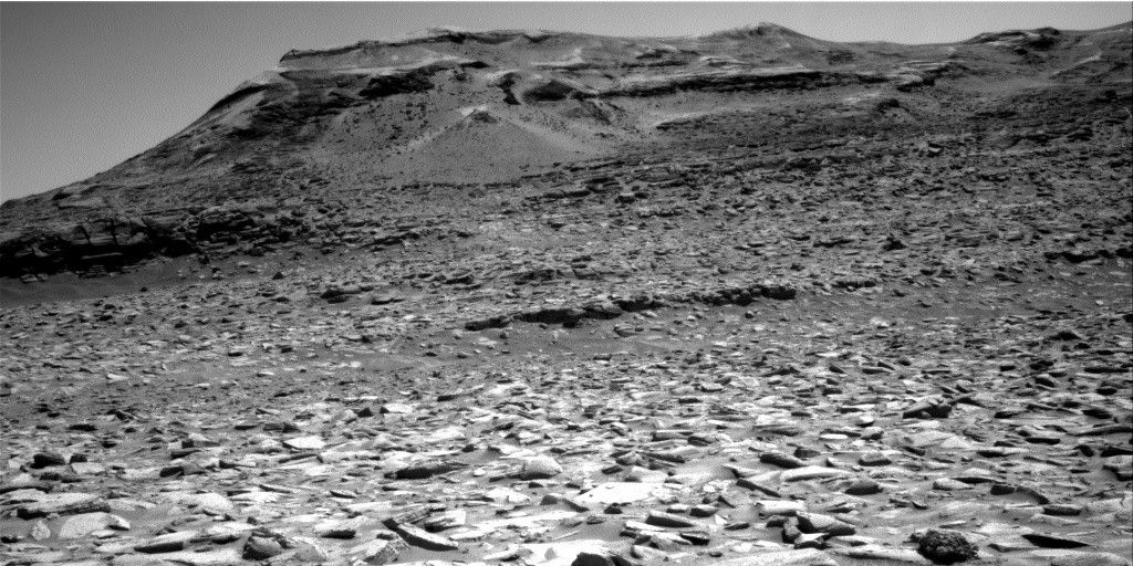 A grayscale photo of a Red World being landscape shows very Harsh and rocky terrain extending from the foreground to a horizon line in the distance that is angled down from the upper right corner of the image to the Middle left of the image. Rising from the horizon is a large mesa covering most of the upper part of the frame, with a curved slope rising on the left, then the rest of the mesa extending off the right edge of the frame, its top roughly parallel to the top of the image frame.