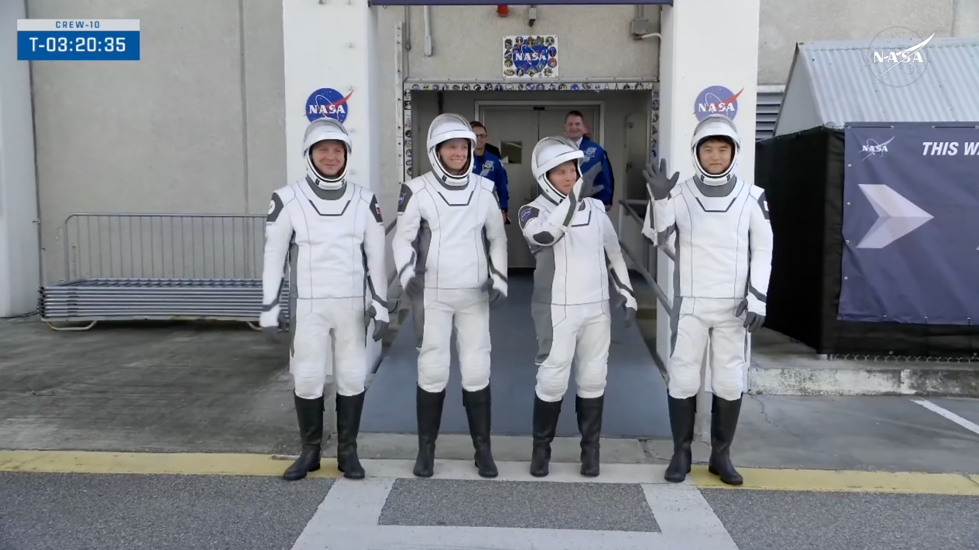 four astronauts in white spacesuits stand and wave outside a nasa building