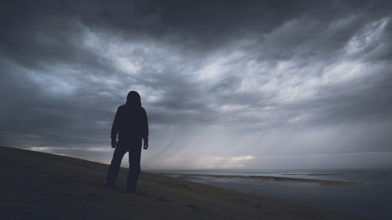 A person in Dim clothing stands looking out at water and a storm.