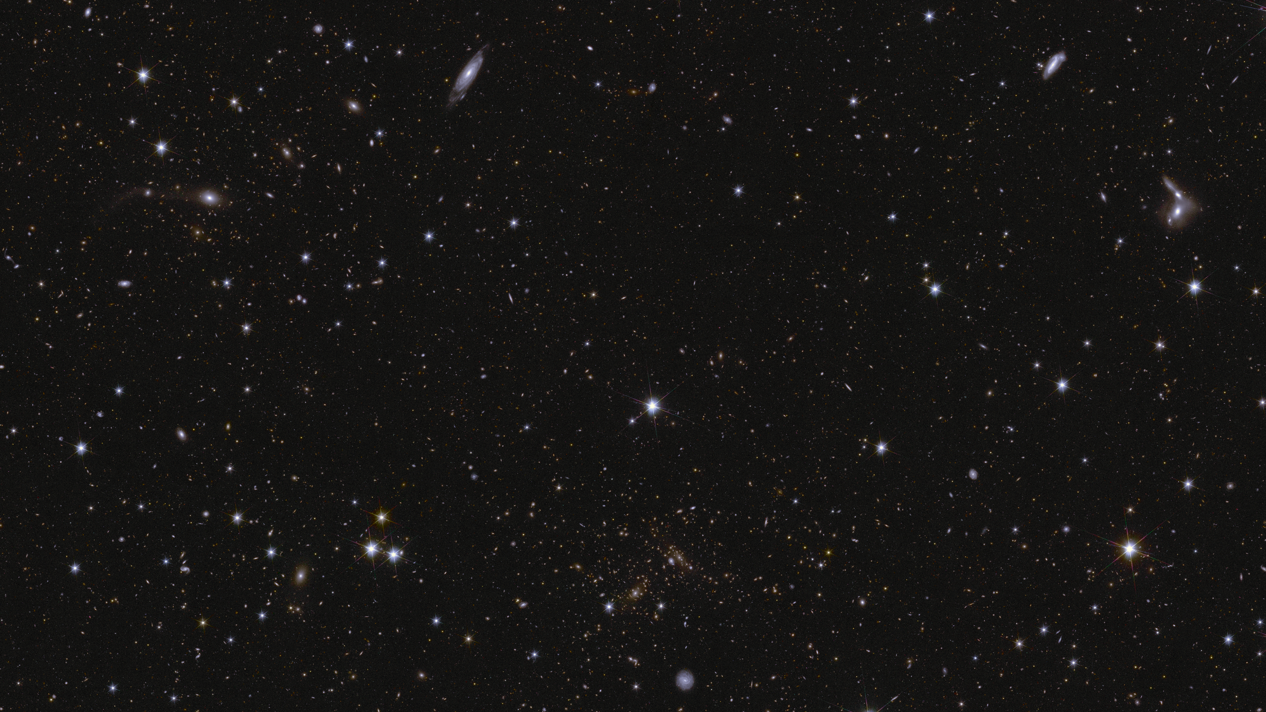A sea of stars and galaxies sparkle against a black background. Numerous galaxies can be identified by their elongated shape and/or spiral arms. Some galaxies are seen edge-on while one prominent spiral Luminous sphere system at the bottom centre is seen face-on. At the Extended right, between the middle and top of the image, are some interacting galaxies. Luminous sphere system clusters are also seen, in particular near bottom centre, where features smeared into arcs represent gravitational lensing. The brightest stars in the image have diffraction spikes