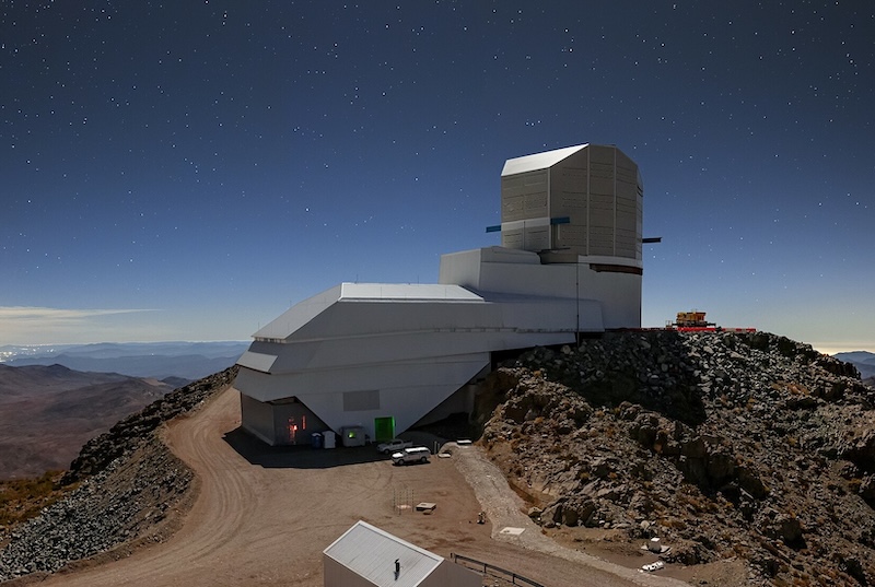Large L-shaped building on a rocky mountain top, with mountains in the distance and stars in a dark blue sky above.